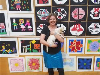 A smiling brunette female holding a dog in front of a panel of 25 various student art pictures.