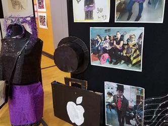 student fashion design project on a mannequin showing a black dress with a purple skirt with a black purse in front of a panel showing the design process and another project with a black hat, a black briefcase with the Apple logo and a blue and white pattern vest with a red and blue striped tie. 
